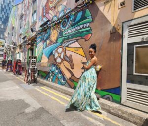 A woman in a flowing green and blue dress poses on a bustling street with vibrant, intricate murals on the walls around her.