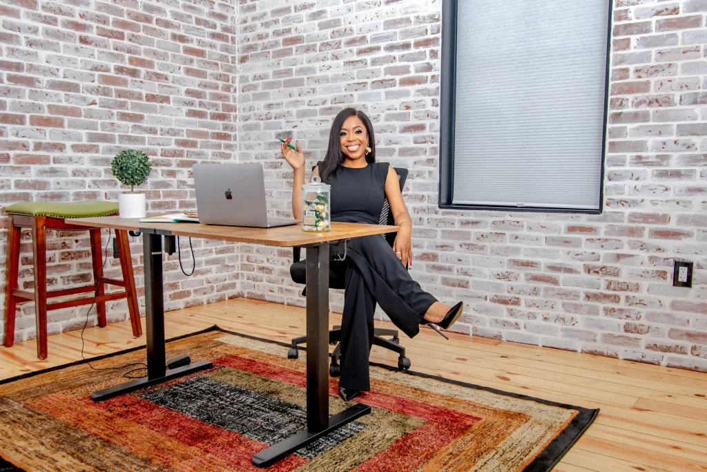 A woman sits at an office desk, smiling and holding a pen. She is in a room with brick walls, a window with a closed blind, and a colorful rug on the floor.