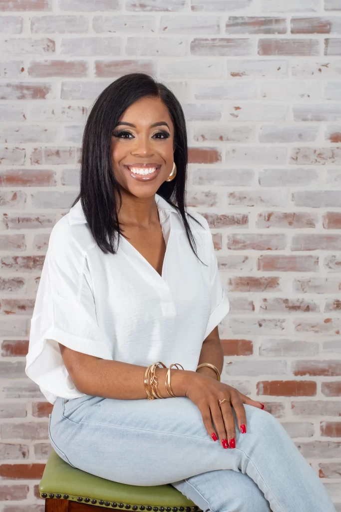A woman with straight black hair is sitting on a stool in front of a white brick wall, smiling. She is wearing a white blouse and blue jeans, and she has red nail polish and gold bracelets.