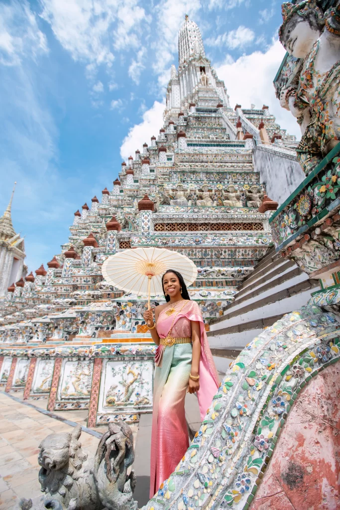 A woman in a traditional dress holds a parasol while standing near a decorated temple in Wat Arun, Thailand. The temple has intricate detailing and a tall spire under a partly cloudy sky.