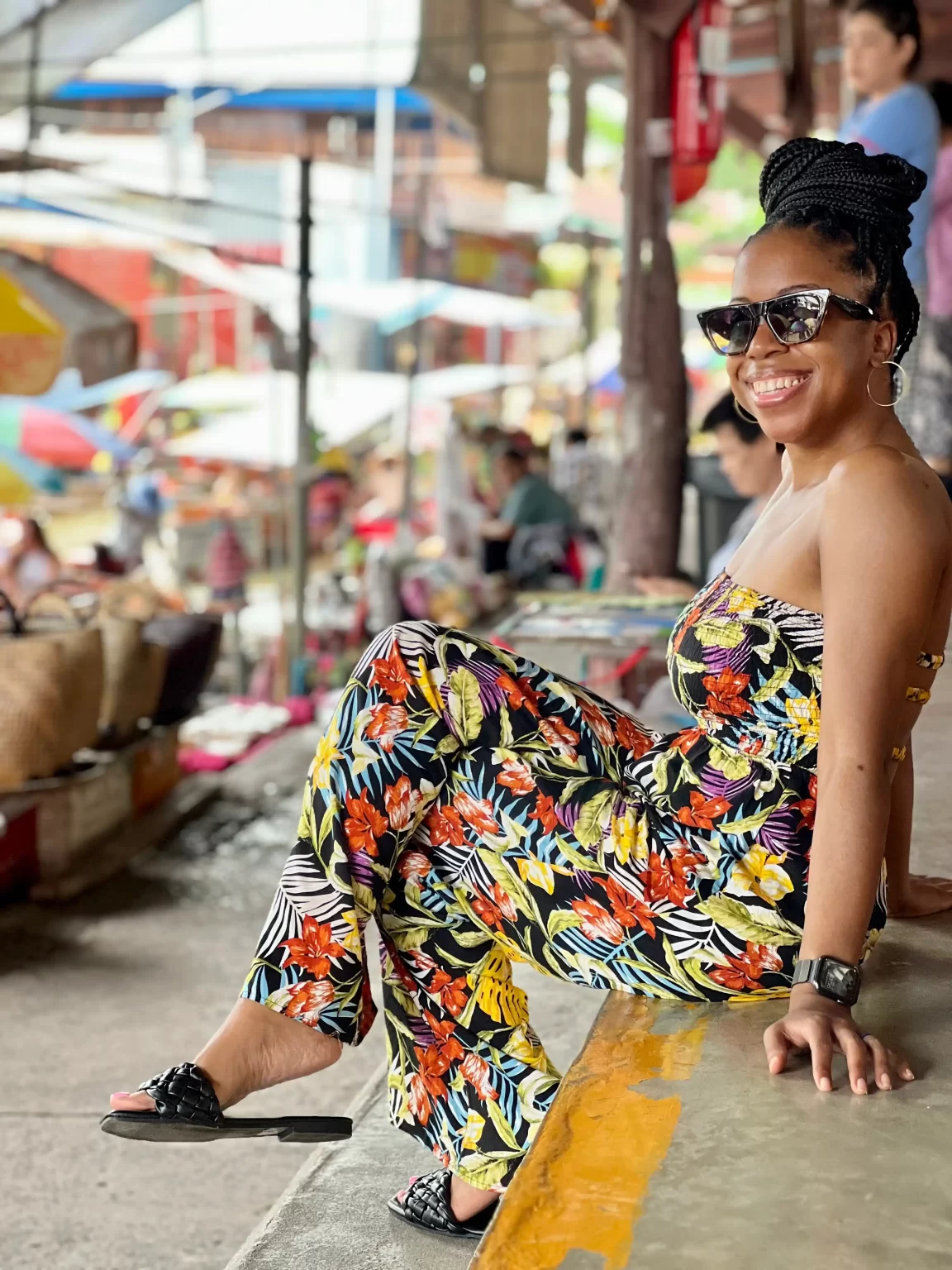 A woman in a floral jumpsuit and sunglasses sits on a ledge, smiling in an outdoor market with colorful umbrellas and various people in the background.