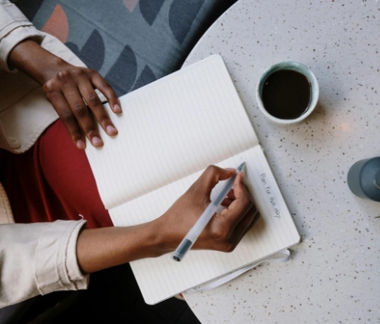 A person writes in an open notebook on a round table with a cup of coffee nearby.