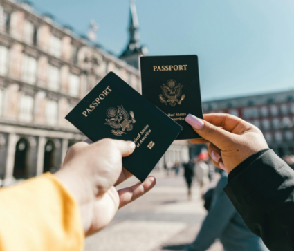Two people are holding United States passports in front of an ornate building in a sunny, outdoor setting.