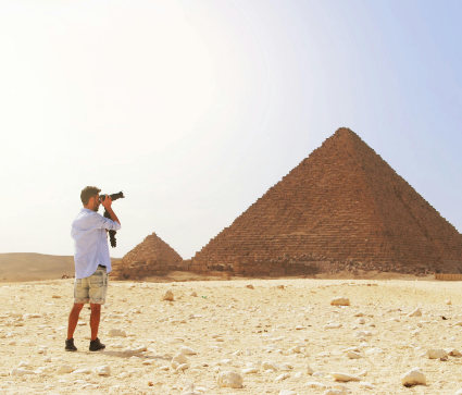 A person in shorts and a light shirt takes photos with a camera near an ancient pyramid in a desert landscape.
