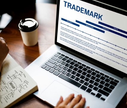 A person is using a laptop displaying the word "TRADEMARK" and a timeline. The person is also writing in a notebook with a cup of coffee nearby.