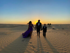A woman in a purple gown and two men in black suits walk hand in hand across a desert at sunrise or sunset.