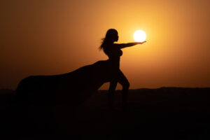 Silhouette of a person with flowing fabric reaching out towards the setting sun, creating an illusion of holding the sun, against an orange sky.
