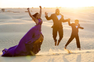 Three people in formal attire jump joyfully in a desert at sunset. A woman in a purple gown, a man in a suit, and a child in a suit are pictured against sand dunes.