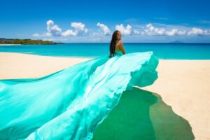 A woman in a flowing turquoise dress stands on a sandy beach with clear turquoise waters and a blue sky with sparse clouds in the background.