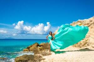 A person in a flowing turquoise dress poses on a rocky shore with the ocean and a bright blue sky in the background.