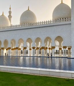 A grand mosque with white domes, intricate arches, golden details, and a reflecting pool in the foreground.