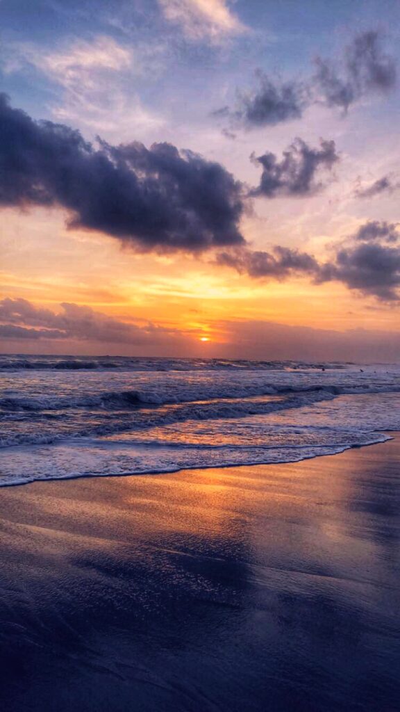 A serene beach sunset with a sky painted in shades of orange and blue. Waves gently roll onto the wet sand, reflecting the colors of the sky.