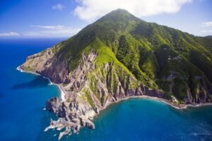 Aerial view of a green-covered mountain with steep cliffs meeting a clear blue sea. Rocks and rugged terrain are visible along the shoreline.