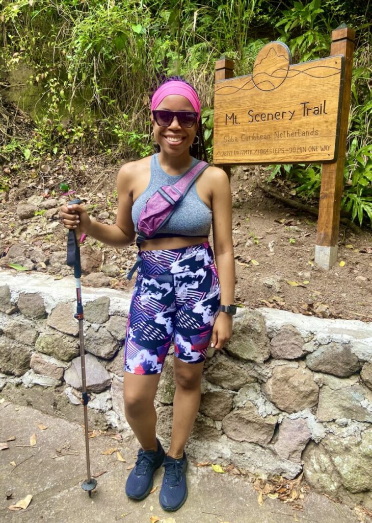 A person stands smiling on a hiking trail, holding a walking stick. They are in front of a sign that reads "Mt. Scenery Trail, Saba Caribbean Netherlands." They wear athletic attire and sunglasses.