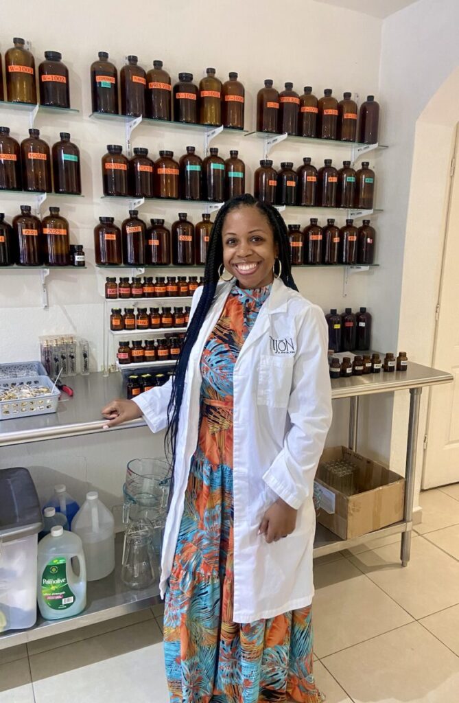 A woman in a floral dress and lab coat stands in a laboratory, smiling. Shelves behind her display multiple brown bottles.