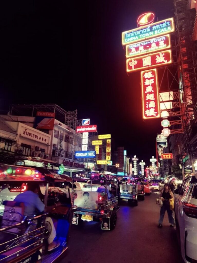 Night view of a busy Chinatown street with illuminated signs in various languages and tuk-tuks navigating the road filled with people.