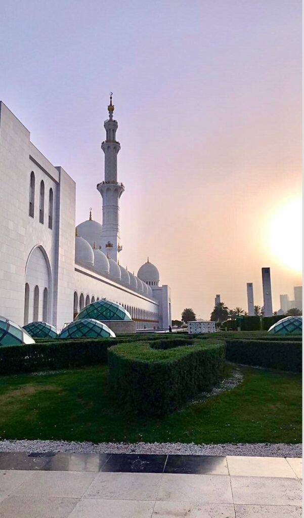 A large white mosque with multiple domes and a tall minaret is set against a hazy sunset sky. Green shrubs and gardens surround the foreground.