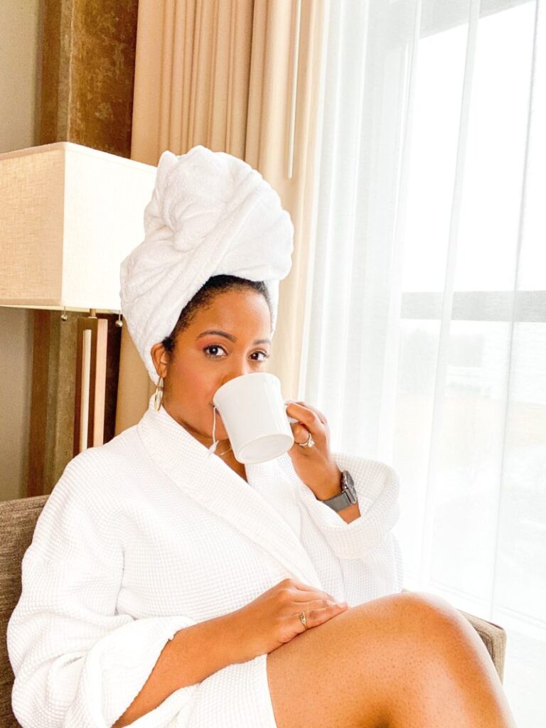 A person wearing a white bathrobe and turban drinks from a white mug while sitting by a window.