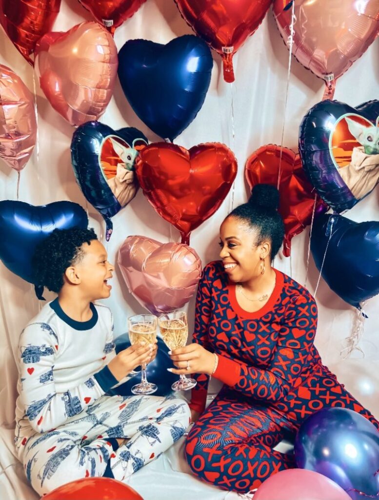 A child and an adult wearing pajamas sit on the floor, clinking glasses with balloons shaped like hearts and stars in the background.