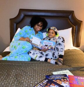 Two people in pajamas sit on a bed; one is reading a book titled "Civil Rights Then and Now" to the other. Several books are spread out at the foot of the bed.