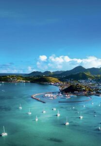 Aerial view of a coastal town with marina, sailboats anchored in a clear blue sea, hills in the background, and a partly cloudy sky above.